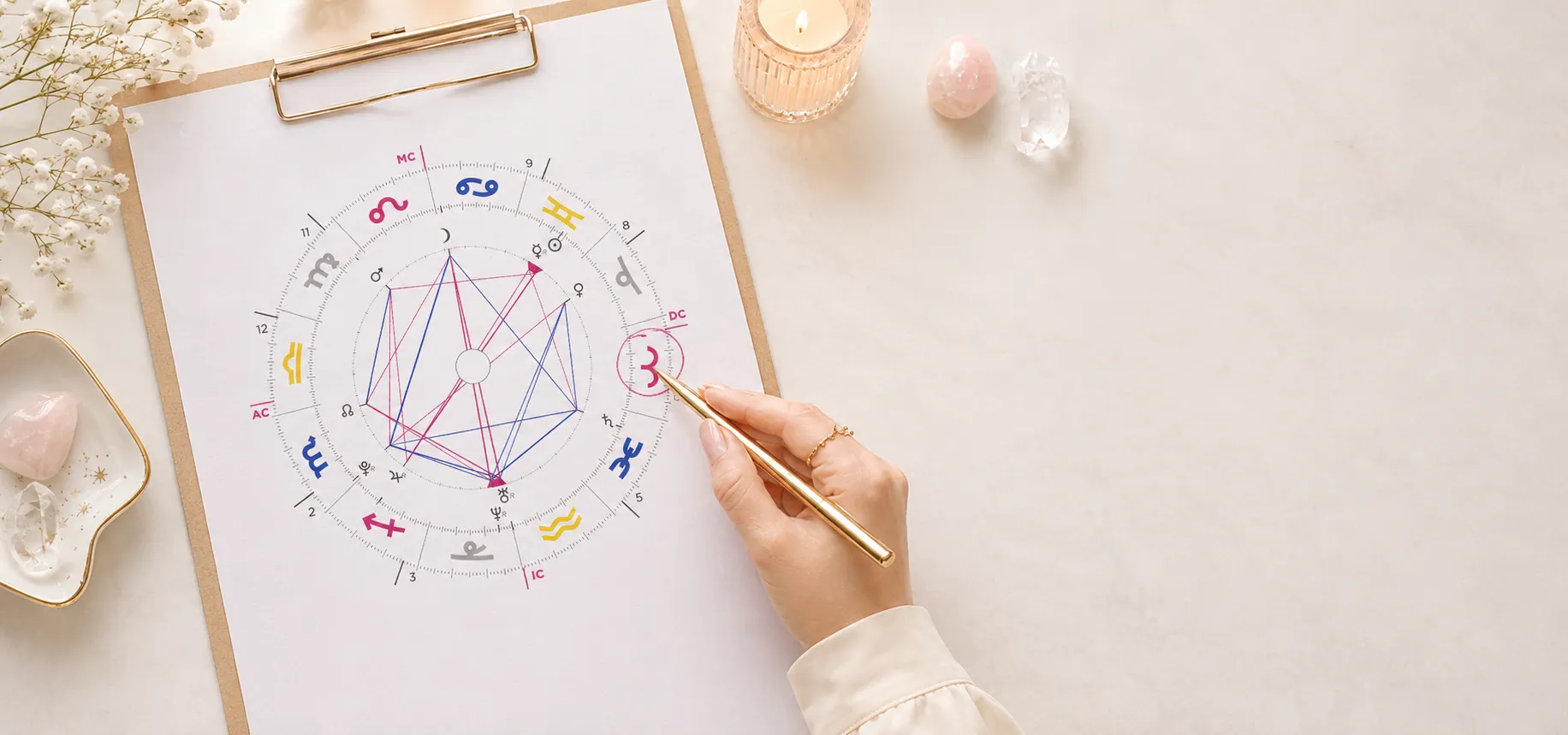 Mapa astral impresso em uma prancheta, com aspectos traçados em rosa e azul, ao lado de uma mão segurando caneta dourada, cristais e uma vela acesa.
